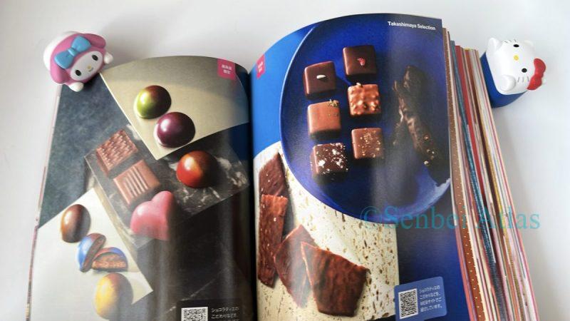 The 120-page event booklet for Amour du Chocolat at Shinjuku Takashimaya Valentine's Day event. The open pages feature colorful photos of chocolates, with My Melody (マイメロディ) peeking from behind the booklet, while Hello Kitty (ハローキティ) stands next to it with her arms raised, looking up