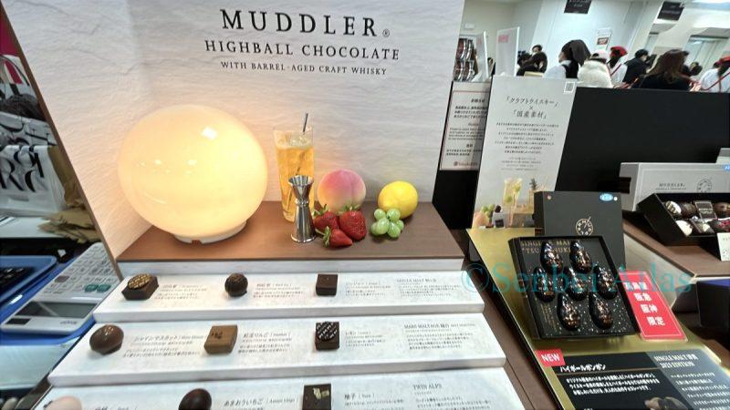 A display of MUDDLER (マドラー) chocolates at the Amour du Chocolat event in Shinjuku Takashimaya. The booth features a bar-themed design  with subdued lighting, cocktail glasses, and imitation fruit to represent their highball-themed chocolates.