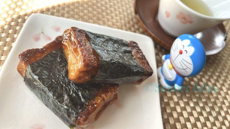 Two pieces of toasted Isobeyaki (Japanese rice cakes wrapped in nori seaweed) served on a plate, with a smiling Doraemon figure watching over them and a Japanese teacup in the background. A cozy, traditional snack scene.