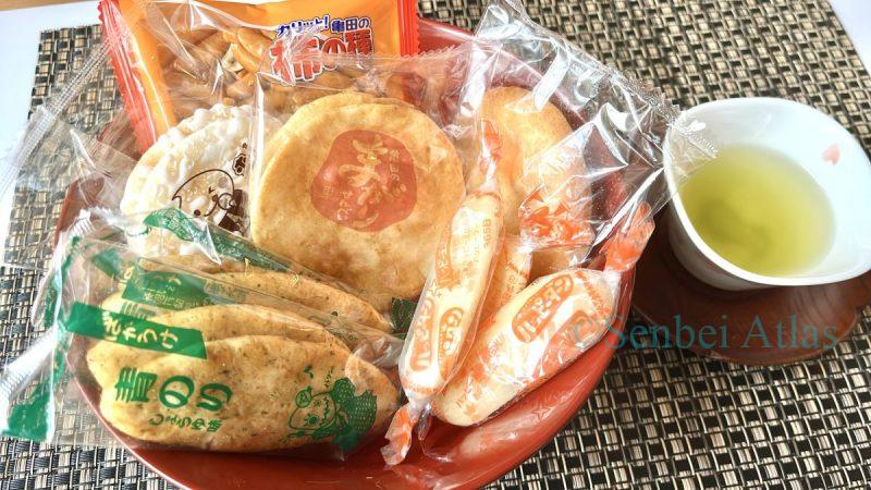 Various types of Japanese rice crackers (Senbei) in their packages: Bakauke (ばかうけ), Teshioya (手塩屋), Magari Senbei (まがりせんべい), Happy Turn (ハッピーターン), Yuki no Yado (雪の宿), and Kaki-no-Tane (柿の種) served with green tea.