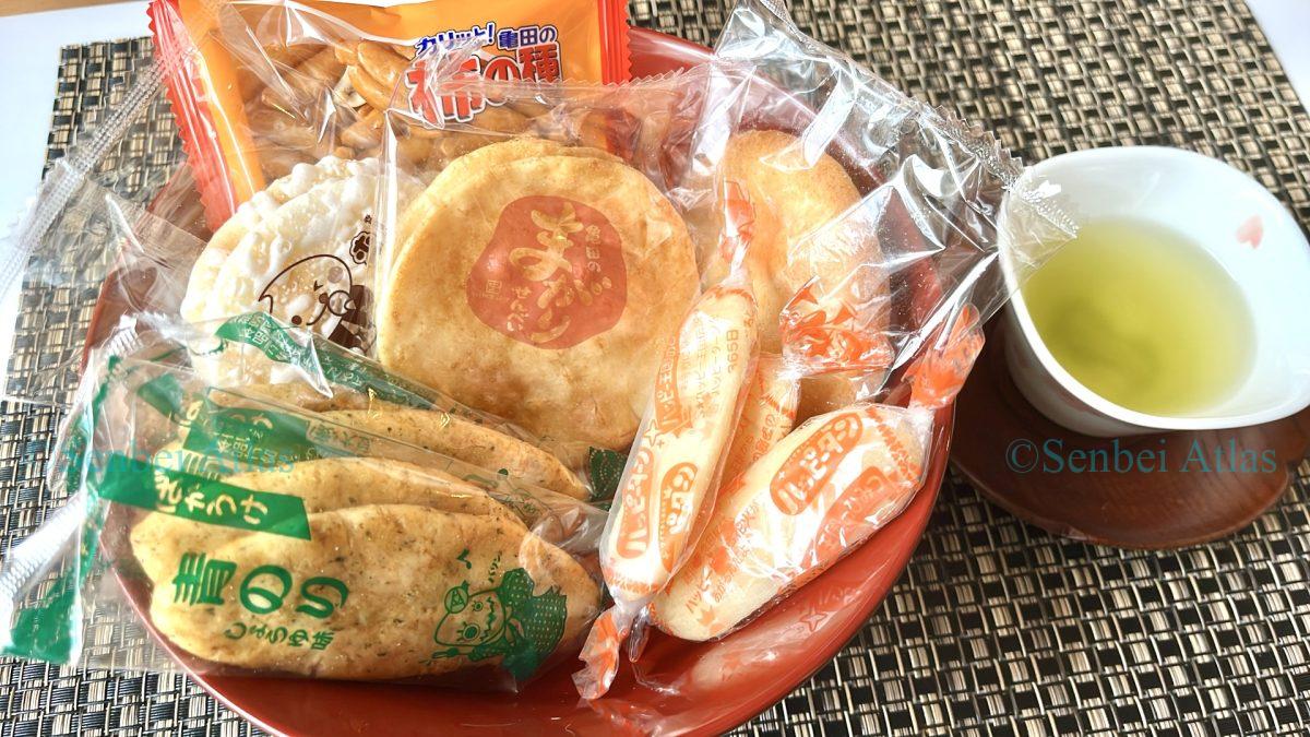 Various types of Japanese rice crackers (Senbei) in their packages: Bakauke (ばかうけ), Teshioya (手塩屋), Magari Senbei (まがりせんべい), Happy Turn (ハッピーターン), Yuki no Yado (雪の宿), and Kaki-no-Tane (柿の種) served with green tea.