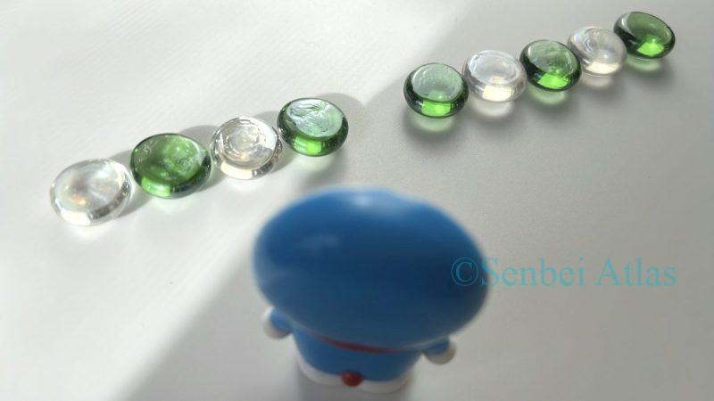 A row of green and white glass beads with a gap, featuring Doraemon in the shadows looking at the missing piece with light shining from the top left.