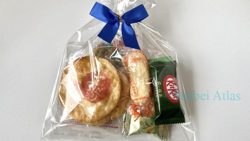 A transparent gift bag tied with a blue ribbon, containing Matcha Kit Kat (抹茶キットカット), Meltykiss Matcha (メルティーキッス抹茶), Happy Turn (ハッピーターン), Magari Senbei (まがりせんべい), and Bakauke (ばかうけ).