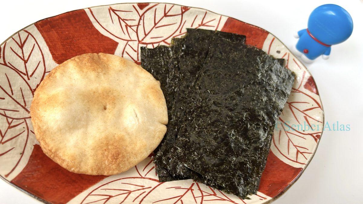 A Doraemon (ドラえもん) figure turning its back on a plate with separated Japanese rice crackers and seaweed. This illustration symbolizes the mystery of the disappearing Norimaki Senbei (のりまきせんべい).