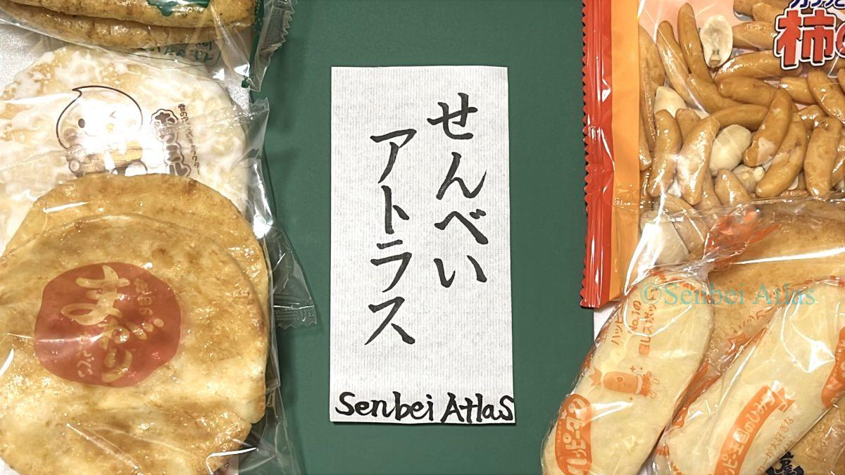 A photograph of hand-brushed Japanese calligraphy reading "Senbei Atlas (せんべいアトラス)" surrounded by various real Japanese rice crackers.