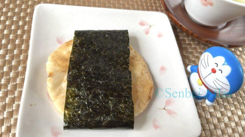 A crispy Magari Senbei (Japanese rice cracker) wrapped with premium Ariake nori seaweed. In the background, a Doraemon figure and a Japanese teacup are visible.