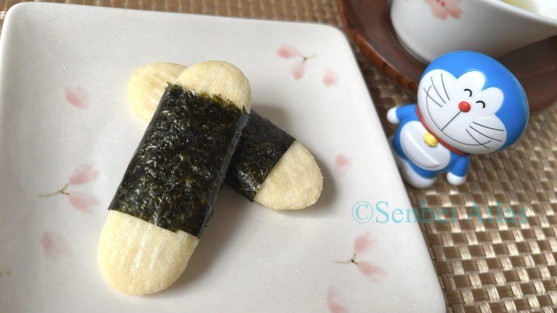 The unique sweet and salty Happy Turn senbei (Japanese rice cracker) wrapped in seaweed. A Japanese teacup and Doraemon figure stand behind the plate.