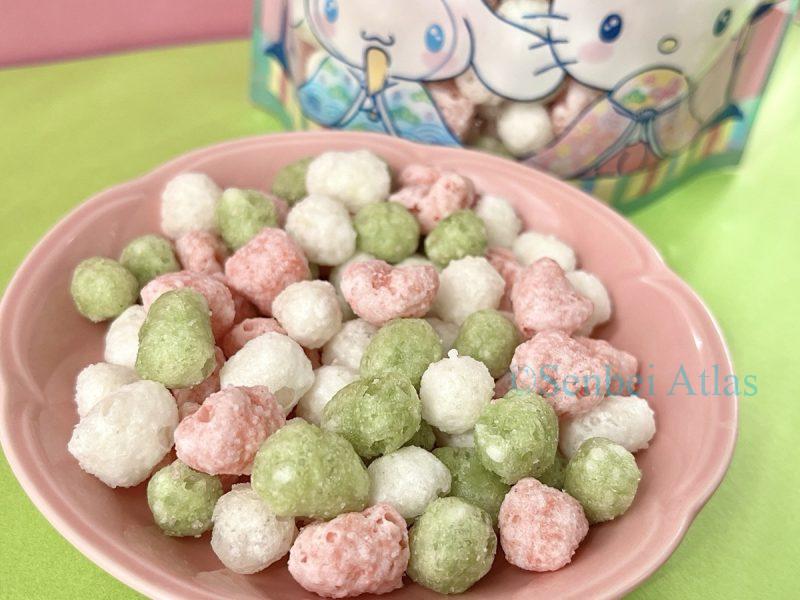 Bite-sized pink, white, and green Hina-arale (ひなあられ) on a plate, with a package in the background showing Cinnamoroll (シナモロール) dressed as an Obina (prince) and Hello Kitty (ハローキティ) as a Mebina (princess).