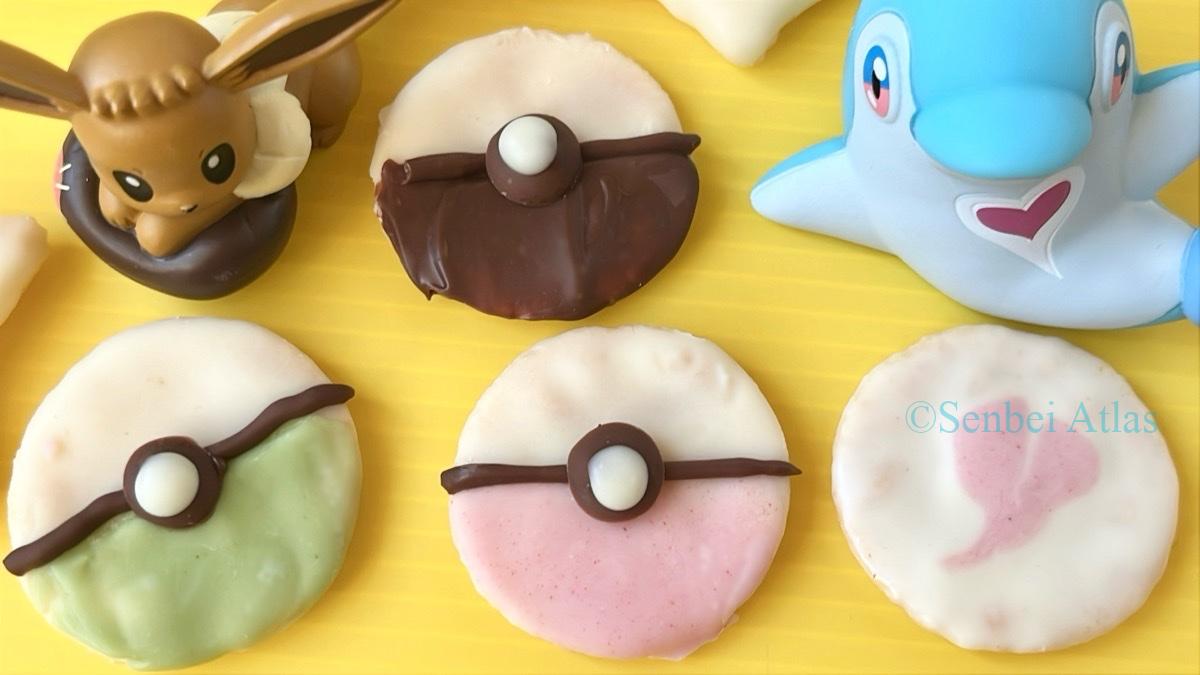 Three colorful chocolate senbei (せんべい) shaped like Poké Balls (ポケボール) from Pokémon (ポケモン), a white chocolate senbei with a pink heart, and figures of Eevee (イーブイ) and Palafin (イルカマン) in the back.