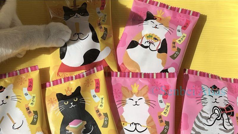 A white cat paw gently touching one of the six Kaldi (カルディ) Hinya-arare (ひにゃあられ) packages.