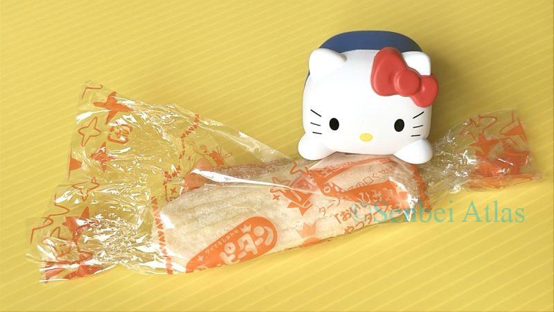 A Hello Kitty figure sitting on top of an individually wrapped Happiturn (ハッピーターン) rice cracker that is partially opened.